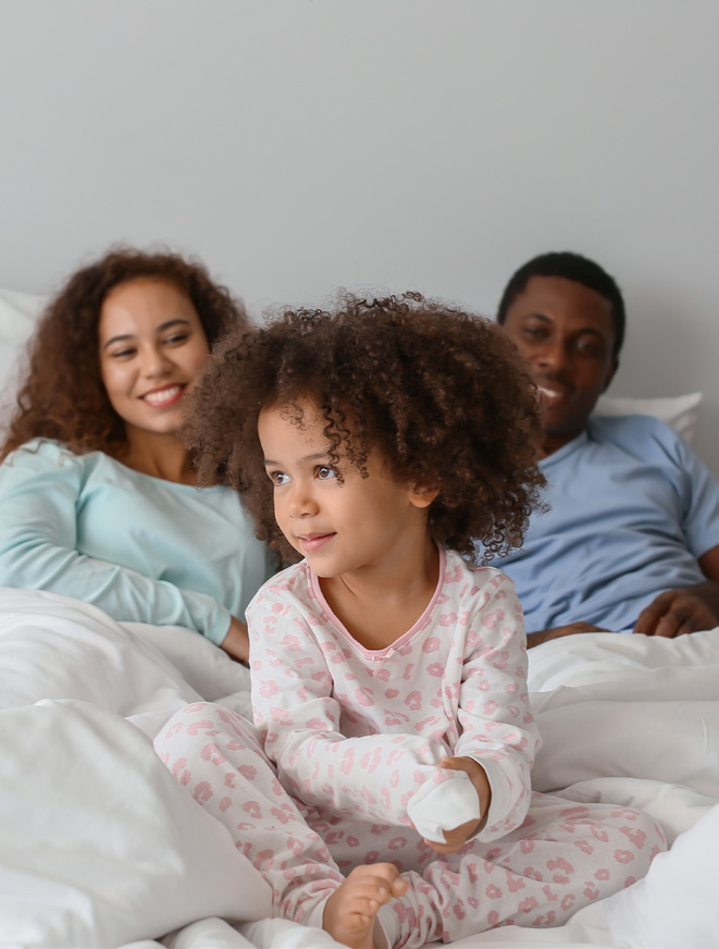 A joyful family morning in bed: a child in pink pajamas sits playfully at the front, with two smiling adults behind in a cozy, relaxed atmosphere.
