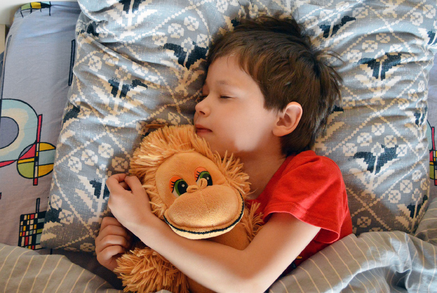 A child in a red shirt peacefully sleeps in bed, hugging a plush monkey toy. The bedding features geometric patterns, conveying warmth and comfort.