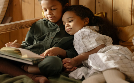 Two children sit closely together reading a book by a wooden wall, bathed in warm sunlight. The scene is cozy and peaceful, conveying a sense of comfort.