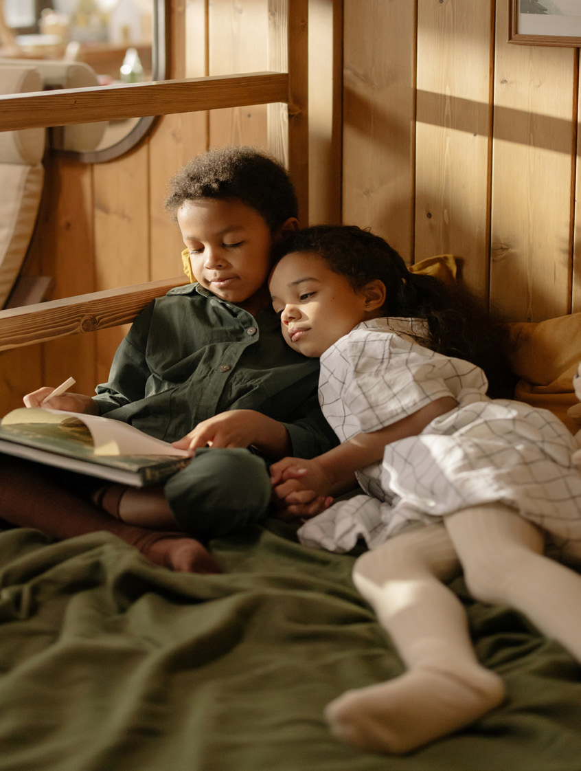Two children sit closely together reading a book by a wooden wall, bathed in warm sunlight. The scene is cozy and peaceful, conveying a sense of comfort.