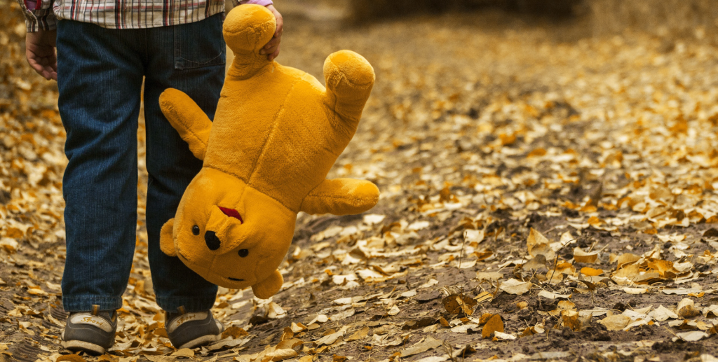 A child in jeans and sneakers walks on a leaf-covered path, holding a large, plush bear upside down, evoking a sense of nostalgia and warmth.