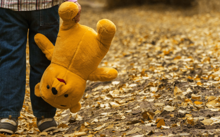 A child in jeans and sneakers walks on a leaf-covered path, holding a large, plush bear upside down, evoking a sense of nostalgia and warmth.
