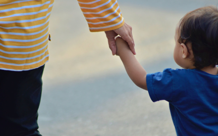 A child in a blue shirt holds hands with an adult in a yellow striped top, walking together on a sunny day. The mood is tender and reassuring.