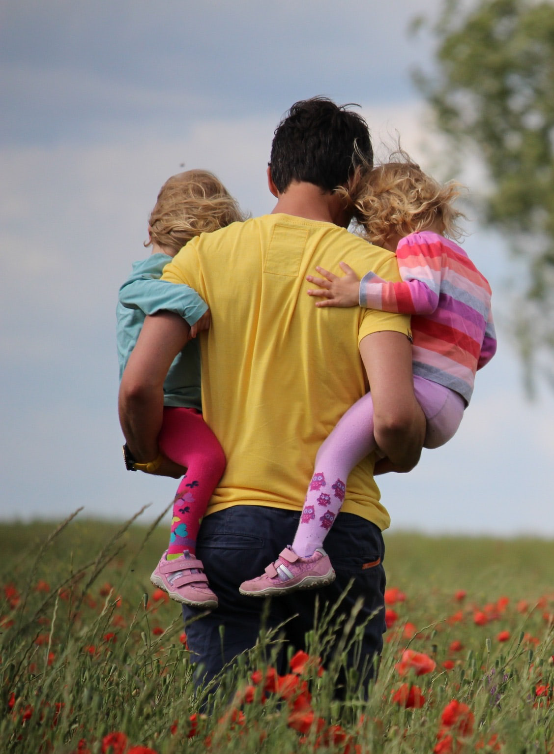 A person in a yellow shirt carries two children in colorful outfits through a field of red flowers. The scene conveys warmth and familial love.