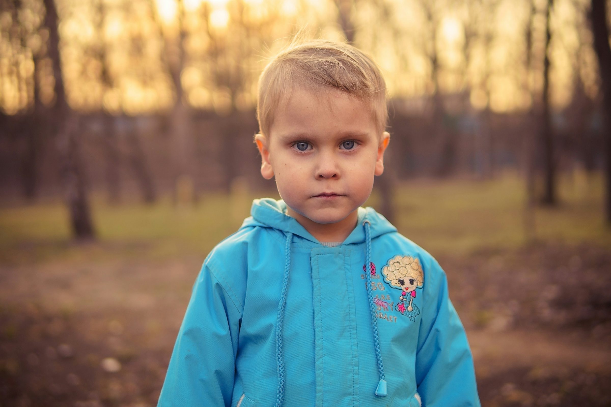 A serious-looking child in a blue jacket stands in a blurred forest at sunset, creating a warm, peaceful, yet contemplative atmosphere.