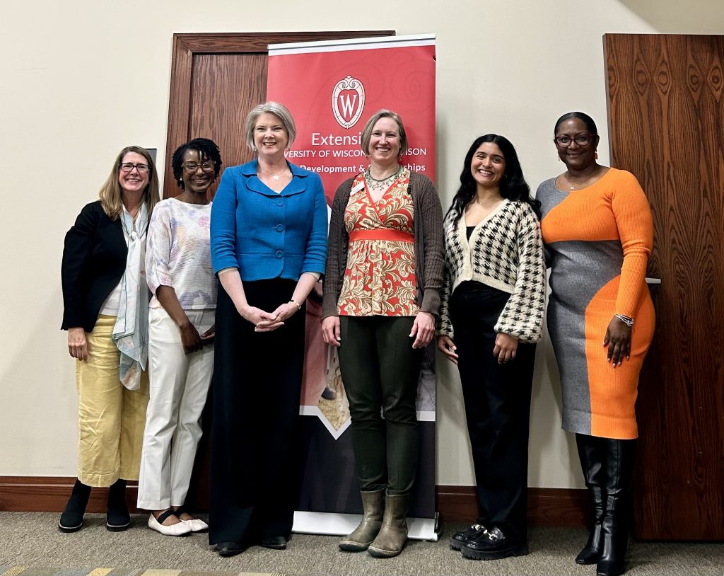 Six women in business clothes stand smiling at a camera against a cream-colored wall with dark brown wood doors. A red sign behind them reads: Extension University of Wisconsin–Madison Human Development & Relationships Institute.