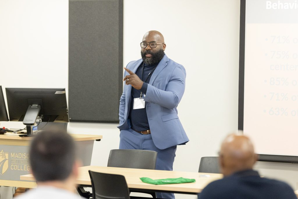 A man in a blue suit with a beard and glasses presents at the front of a classroom
