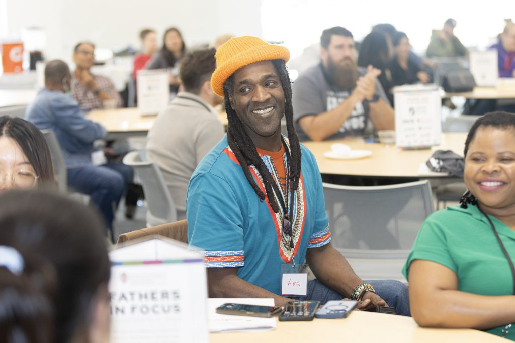 A man in a yellow hat and blue shirt smiles at someone off camera while a woman in the green shirt does the same.