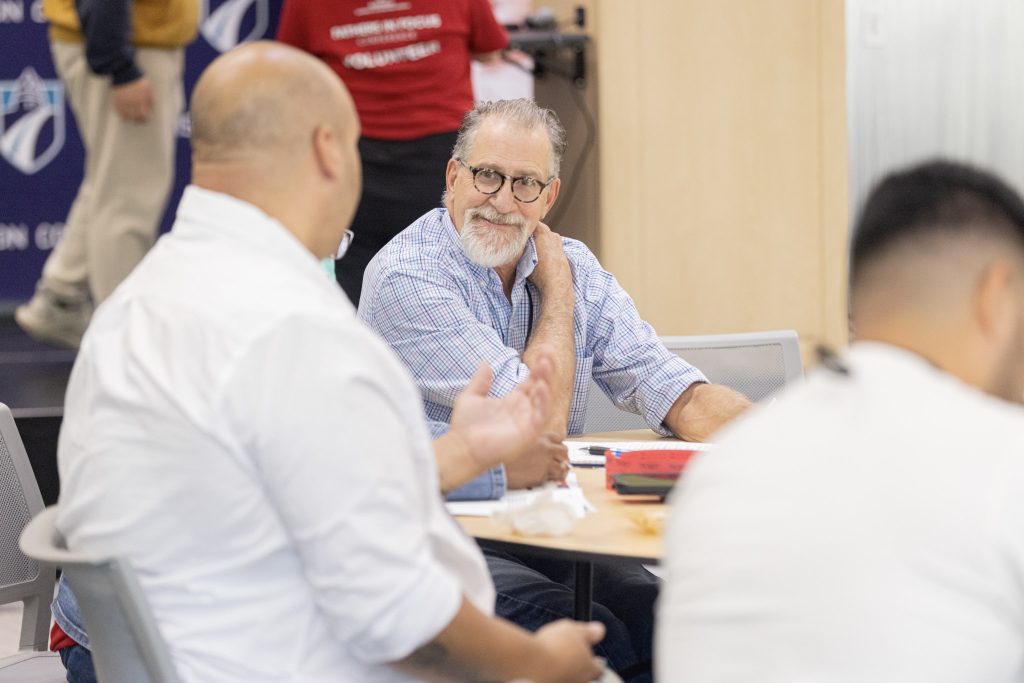 A man with grey hair and glasses smiles at another man while both sit at a table