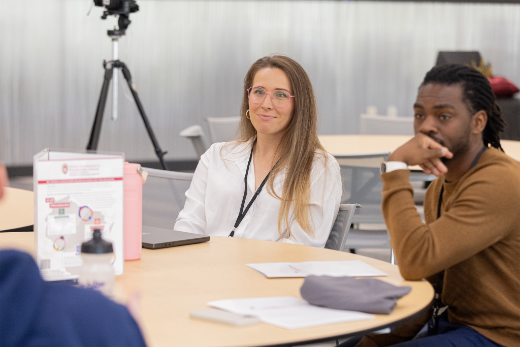 A woman in glasses and a man wearing a watch are seated at a table and look at someone talking off camera
